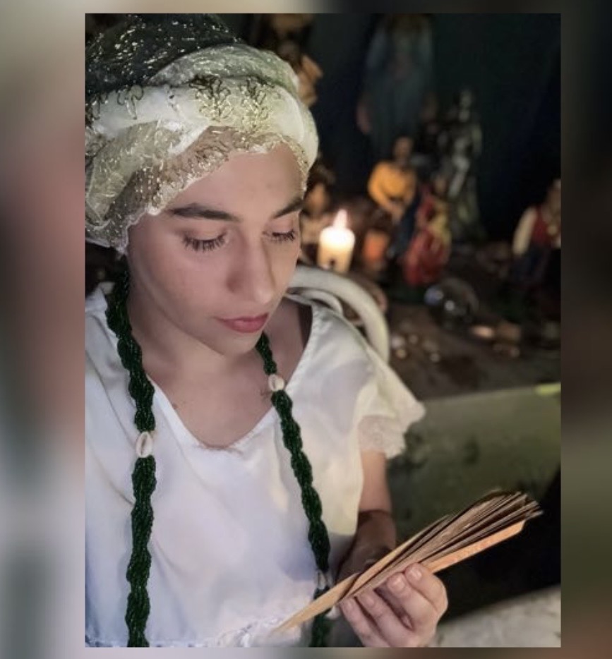 Mãe Glória em atendimento espiritual, com vestes brancas e guias, diante de um altar com vela acesa
